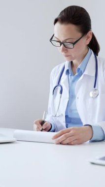 Orta yaşlı kadın doktor, sağlık evraklarını tamamlıyor, dizüstü bilgisayarın yanına yerleştirilmiş bir pano ve steteskop, pencerenin yanında iyi aydınlatılmış klinik çalışma alanı. İlaç ve sağlık hizmetleri.