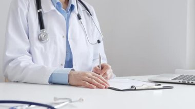 Sağlık evraklarını tamamlayan kadın doktor, dizüstü bilgisayarın yanına yerleştirilmiş bir pano ve cam kenarında iyi aydınlatılmış bir klinik çalışma alanında stetoskop. İlaç ve sağlık hizmetleri kavramı.