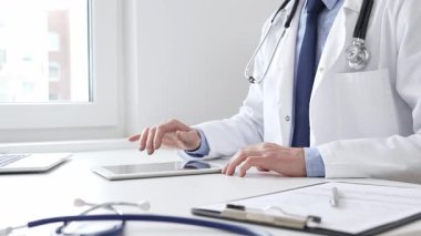 Laboratuvar önlüğü ve steteskop giyen erkek doktor, tıbbi muayenehanede masasında otururken dijital tablet kullanıyor..