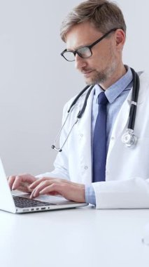 Orta yaşlı doktor özenle bir dizüstü bilgisayarda yazıyor, muhtemelen hasta kayıtlarını güncelliyor ya da araştırma yapıyor, sağlık ve teknolojinin tıbbi ortamdaki kesişimini gösteriyor. İlaç..