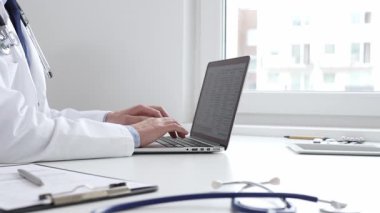 Doktor özenle bir dizüstü bilgisayarda yazıyor, muhtemelen hasta kayıtlarını güncelliyor veya araştırma yapıyor, modern bir tıbbi ortamda sağlık ve teknolojinin kesişimini gösteriyor.