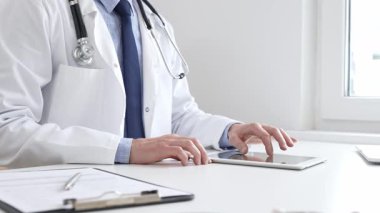 Laboratuvar önlüğü ve steteskop giyen erkek doktor, tıbbi muayenehanede masasında otururken dijital tablet kullanıyor..