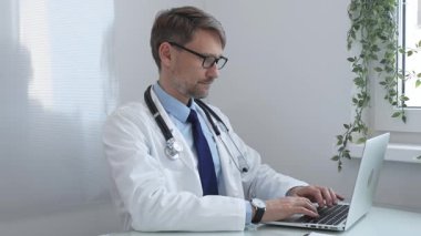 Laboratuvar önlüğü ve steteskop giyen erkek doktor, bir masada oturuyor ve modern bir tıp ofisinde dizüstü bilgisayarda yazıyor, hasta bakımı ve konsültasyona odaklanıyor. İlaç ve sağlık hizmetleri.