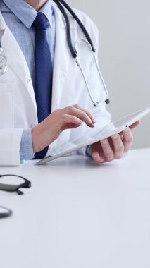 Laboratuvar önlüğü ve steteskop giyen erkek doktor, tıbbi muayenehanede masasında otururken dijital tablet kullanıyor..
