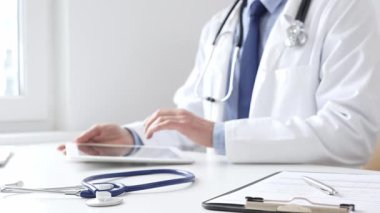 Klinikte dijital tablet kullanan doktor adamın yanındaki masada mavi steteskop duruyor. Tıp ve sağlık hizmetleri kavramı