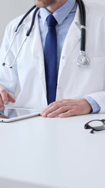 Laboratuvar önlüğü ve steteskop giyen erkek doktor, tıbbi muayenehanede masasında otururken dijital tablet kullanıyor..