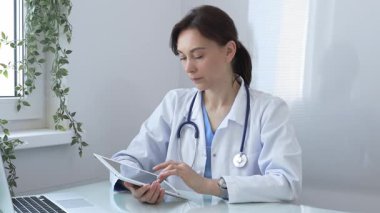 Laboratuvar önlüğü ve steteskop giyen kendine güvenen kadın doktor tıbbi ofiste masasında otururken dijital tablet kullanıyor, hasta bilgilerine bakıyor ya da çevrimiçi danışmanlık yapıyor. Tıp konsepti.