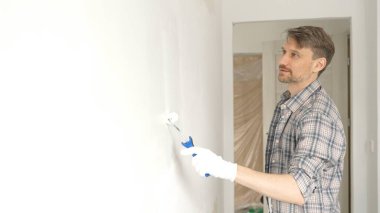 Kareli gömlek giyen profesyonel erkek ressam, apartman duvarına beyaz boya sürüyor. Kapsamlı ev tadilatı sırasında düzenli darbeler alarak iç mekanı değiştiriyor..