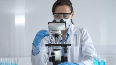 Laboratuvar önlüğü ve mavi eldivenli bir kadın bilim adamı steril bir laboratuvarda mikroskop slaytını inceliyor ve test tüpündeki mavi örnekleri kullanarak araştırma yapıyor..