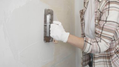 Eldiven giyen kimliği belirsiz kadın inşaat işçisi duvara sıva uygulamak için macun bıçağı kullanıyor. Ev yenileme projesi sırasında yüzeyi düzleştiriyor.