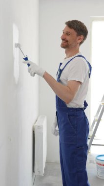 Koruyucu beyaz eldiven ve mavi iş tulumu giyen profesyonel bir erkek ressam. Ev yenileme projesi sırasında el değmemiş iç duvarlara ruloyla boya sürüyor..