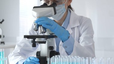 Koruyucu ekipman kullanan bir bilim adamı mikroskop merceğini ayarlar ve steril bir laboratuvar ortamında tıbbi araştırma örneklerini doğru bir şekilde analiz eder..