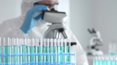 Laboratuvar ortamında araştırma yaparken mikroskop kullanarak koruyucu kıyafet giyen bir tıp bilimcisi. Test tüplerini mavi sıvıyla kapatın..