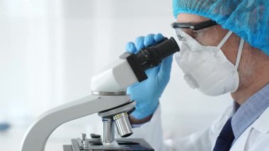 Koruyucu ekipman takan erkek bilim adamı mikroskop kaydırağını inceliyor, steril laboratuvar ortamında araştırma örneklerini analiz ediyor. Sağlık ve bilim konsepti.