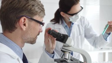 Bilimsel uzmanlar mikroskopla numuneleri inceliyor. Araştırma sırasında her ikisi de beyaz laboratuvar önlüğü giymiş koruyucu maske takan laboratuvar asistanı. Tıp ve Bilim.