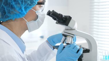 Laboratuvarda mikroskopla araştırma örneği inceleyen kadın bir bilim adamı tıbbi araştırma için beyaz ve mavi laboratuvar malzemesi giyiyor. Tıp, bilim konsepti.