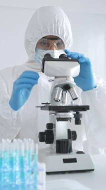 Koruyucu giysi giyen tıp bilimcisi örnekleri incelerken mikroskop kullanıyor steril laboratuvar ortamında araştırma yapıyor. Mavi sıvıyla test tüplerinin yakın plan görüntüsü.