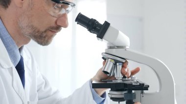 Yetişkin erkek bilim araştırmacısı mikroskopla örnek analiz ediyor, yüksek teknoloji laboratuvar ortamında koruyucu giysiler giyiyor. Sağlık, tıp ve bilim konsepti.
