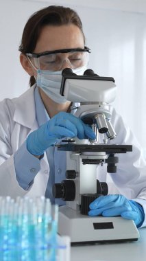 Mikroskop kaydırağını inceleyen kadın bilim adamı, laboratuarda koruyucu giysiler giyiyor, mavi renkli test tüpleriyle farmasötik buluşları araştırıyor..