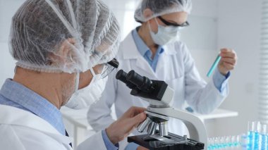 Erkek bilim adamı koruyucu maske takıyor, gözlük takıyor ve mikroskopla çalışıyor. Bu sırada bayan meslektaşı da tableti tutmasına ve laboratuarda notlar almasına yardım ediyor. Tıp ve bilim konsepti.