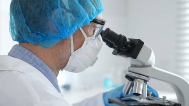 Koruyucu ekipman takan erkek bilim adamı mikroskop kaydırağını inceliyor, steril laboratuvar ortamında araştırma örneklerini analiz ediyor. Sağlık ve bilim konsepti.