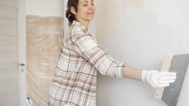 Kadın duvara macun bıçağıyla alçı yapıştırıyor, koruma için eldiven takıyor ve tadilat sırasında bile pürüzsüz ve sorunsuz bir şekilde bitmesini sağlıyor.