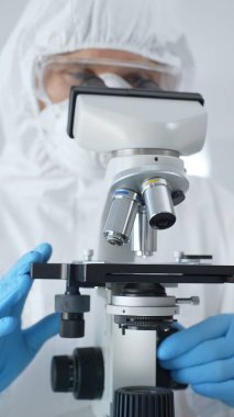 Tehlikeli madde giysisi, maske ve mavi eldiven giyen erkek bir virüs bilimci laboratuvarda virüsleri araştırırken mikroskop altında özenle çalışıyor. Tıp ve bilim konsepti.