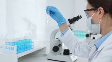 Koruyucu maske takan dişi araştırmacı ve mavi eldiven test tüpünde sıvıyı inceliyor, laboratuvarda mikroskop ve laboratuvar ekipmanlarının yanında oturuyor. Tıp, sağlık ve bilim konsepti.