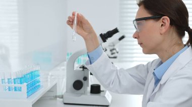 Laboratuvar önlüğü giyen ve koruyucu gözlük takan kadın bilim adamı modern laboratuarda mikroskop ve test tüpleri askısı ile test tüpünü temiz sıvıyla analiz ediyor. Tıp, sağlık ve bilim konsepti