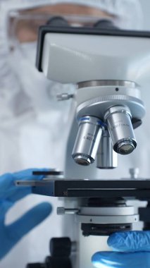 Tehlikeli madde giysisi, maskesi ve eldiveni olan bir tıp virologu bir laboratuvar araştırma virüsünün mikroskobunda özenle çalışıyor. Sağlık ve Bilim Konsepti.