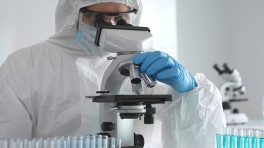 Tehlikeli madde giysisi giyen tıp virolojisti laboratuvarda mikroskopla çalışıyor. Yeni virüsler için potansiyel tedavileri araştırırken mavi sıvı örneklerini inceliyor.