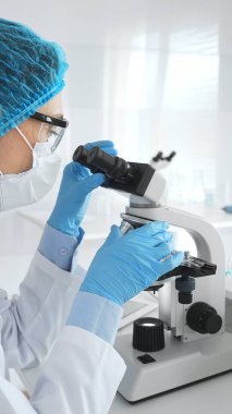 Mavi koruyucu giysiler giyen kadın bilim adamı mikroskop kullanarak laboratuvar ortamında örnekler inceleyerek bilimsel araştırma ve analiz yapıyor. Tıp ve bilim konsepti.