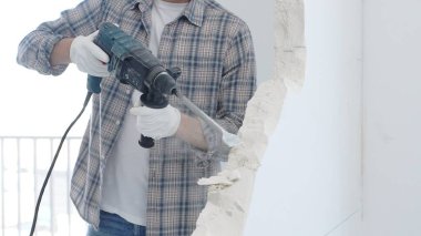 Beyaz duvarı döner çekiçle delen erkek inşaat işçisi gri kareli gömlek ve koruyucu beyaz eldiven giyiyor, yenileme projesi sırasında toz üretiyor, yakın plan