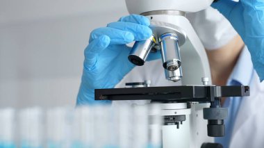 Mikroskobu ayarlayan eldivenli bilim adamlarına yakın çekim yapmak, laboratuvarda araştırma yapmak ve arka planda mavi sıvı içeren test tüpleri kullanmak. Tıp, sağlık ve bilim konsepti.