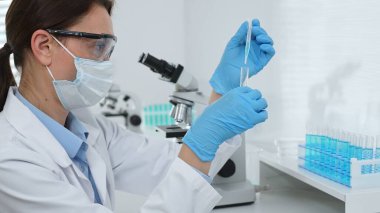 Yüz maskesi takan ve mavi eldiven takan kadın bir bilim adamı modern bir laboratuarda bir deney tüpüne dikkatlice sıvı bırakıyor. Tıp ve bilim konsepti.