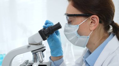 Tıbbi araştırma sırasında steril laboratuvar ortamında çalışırken mikroskop üzerinden numune inceleyen kadın bilim adamı. Tıp, sağlık ve bilim konsepti