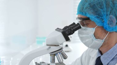 Kıdemli bilim adamı mikroskopla numune inceliyor, steril laboratuvar ortamında tıbbi araştırma yaparken koruyucu giysiler giyiyor. Tıp, sağlık ve bilim konsepti