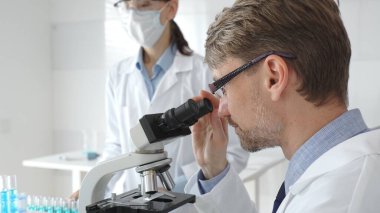 Erkek bir araştırmacı bir örneği mikroskop altında inceliyor, koruyucu maskeli kadın bir laboratuvar teknisyeni çalışma sırasında beyaz laboratuvar önlükleriyle yakınlarda duruyor. Tıp ve Bilim.