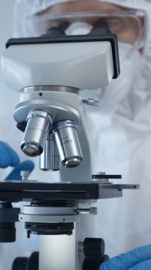 Tehlikeli madde giysisi, maskesi ve eldivenleri olan bir tıp virologu laboratuvar araştırmalarında mikroskop başında özenle çalışıyor. Tıp ve bilim konsepti.