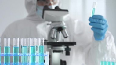 Laboratuvar ortamında araştırma yaparken mikroskop kullanarak koruyucu kıyafet giyen bir tıp bilimcisi. Test tüplerini mavi sıvıyla kapatın..