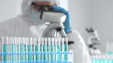 Laboratuvar ortamında araştırma yaparken mikroskop kullanarak koruyucu kıyafet giyen bir tıp bilimcisi. Test tüplerini mavi sıvıyla kapatın..