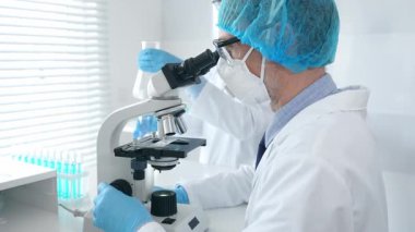 Koruyucu maske takan ve gözlük takan erkek bilim adamı mikroskop kullanarak laboratuvarda araştırma yapıyor ve elinde mataralar olan bir bilim adamı asistanı var. Sağlık ve bilim konsepti.