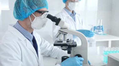 Koruyucu maske takan ve gözlük takan erkek bilim adamı mikroskop kullanarak laboratuvarda araştırma yapıyor ve elinde mataralar olan bir bilim adamı asistanı var. Sağlık ve bilim konsepti.