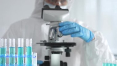 Laboratuvar ortamında araştırma yaparken mikroskop kullanarak koruyucu kıyafet giyen bir tıp bilimcisi. Test tüplerini mavi sıvıyla kapatın..