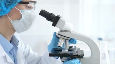 Araştırma örneğini mikroskop üzerinden inceleyen kadın bilim adamı tıbbi araştırma için koruyucu laboratuvar malzemesi giyiyor. Laboratuvar ve bilim konsepti.