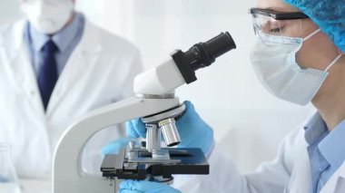 Koruyucu giysili kadın bilim adamı mikroskop altında numuneleri incelerken laboratuvar asistanı profesyonel bir tıbbi araştırmada cam ile ilgileniyor. Tıp ve bilim konsepti.