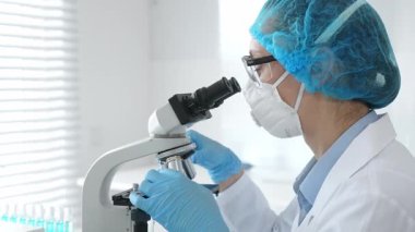 Kadın araştırmacı mikroskop ile numune inceliyor, steril laboratuvar içinde çalışma yerini çevreleyen cam şişelerle tıbbi deney yapıyor. Tıp ve Bilim.