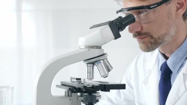 Yetişkin erkek bilim araştırmacısı mikroskopla örnek analiz ediyor, yüksek teknoloji laboratuvar ortamında koruyucu giysiler giyiyor. Sağlık, tıp ve bilim konsepti.