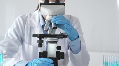 Mikroskobu ayarlayan, laboratuardaki örnekleri inceleyen, yeni ilaç veya kimyasal formülasyon araştırmaları yapan laboratuvar önlüğü giyen ve güvenlik için yüz maskesi takan bilim insanlarının eldiveni kapalı.