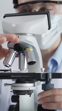 Kadın araştırmacı mikroskop ile numune inceliyor, steril laboratuvar içinde çalışma yerini çevreleyen cam şişelerle tıbbi deney yapıyor. Tıp ve Bilim.